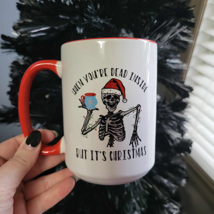 All Decked Out 15oz ceramic mug that says "When you're dead inside but it's Christmas" with a skeleton wrapped in Christmas lights wearing a Santa hat