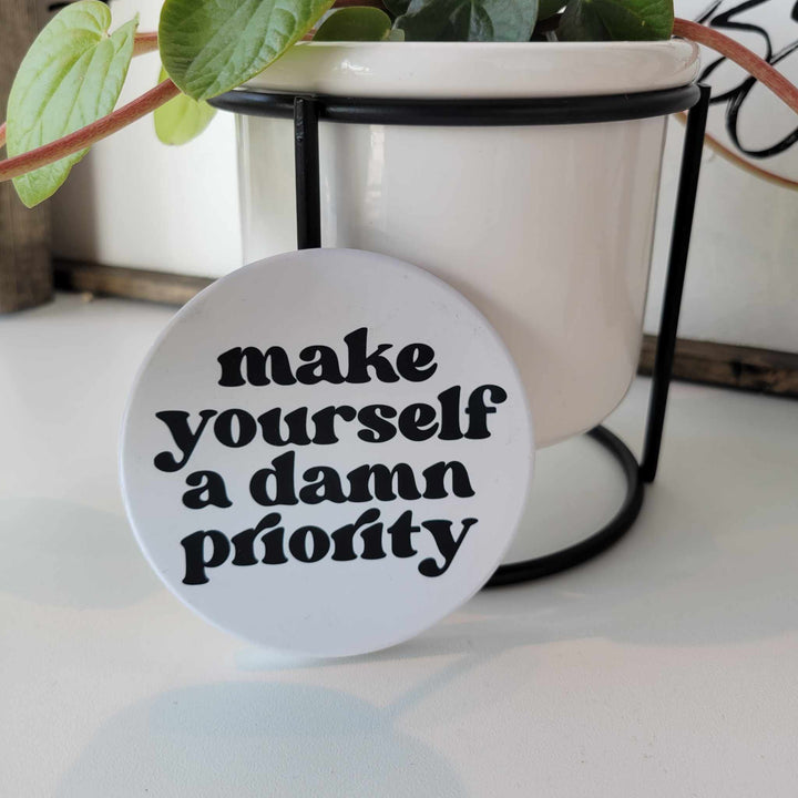 Smooth matte finish ceramic coaster that says "Make yourself a damn priority" in black lettering. Bumper dots on the bottom.